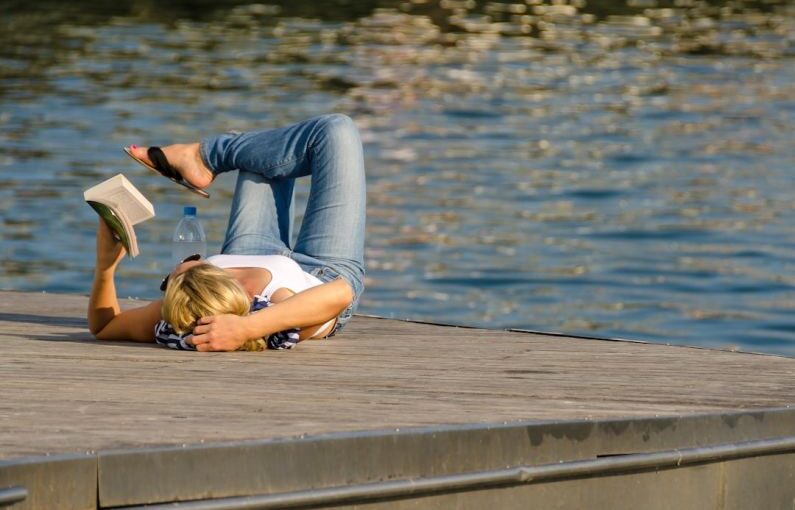Downtime - woman in white tank top and gray pants lying on brown wooden dock during daytime