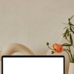 Flat Designs - Netbook with white screen placed on shelf near uneven curved ceramic vases with flowers with green leaves near pipes in light room with white wall